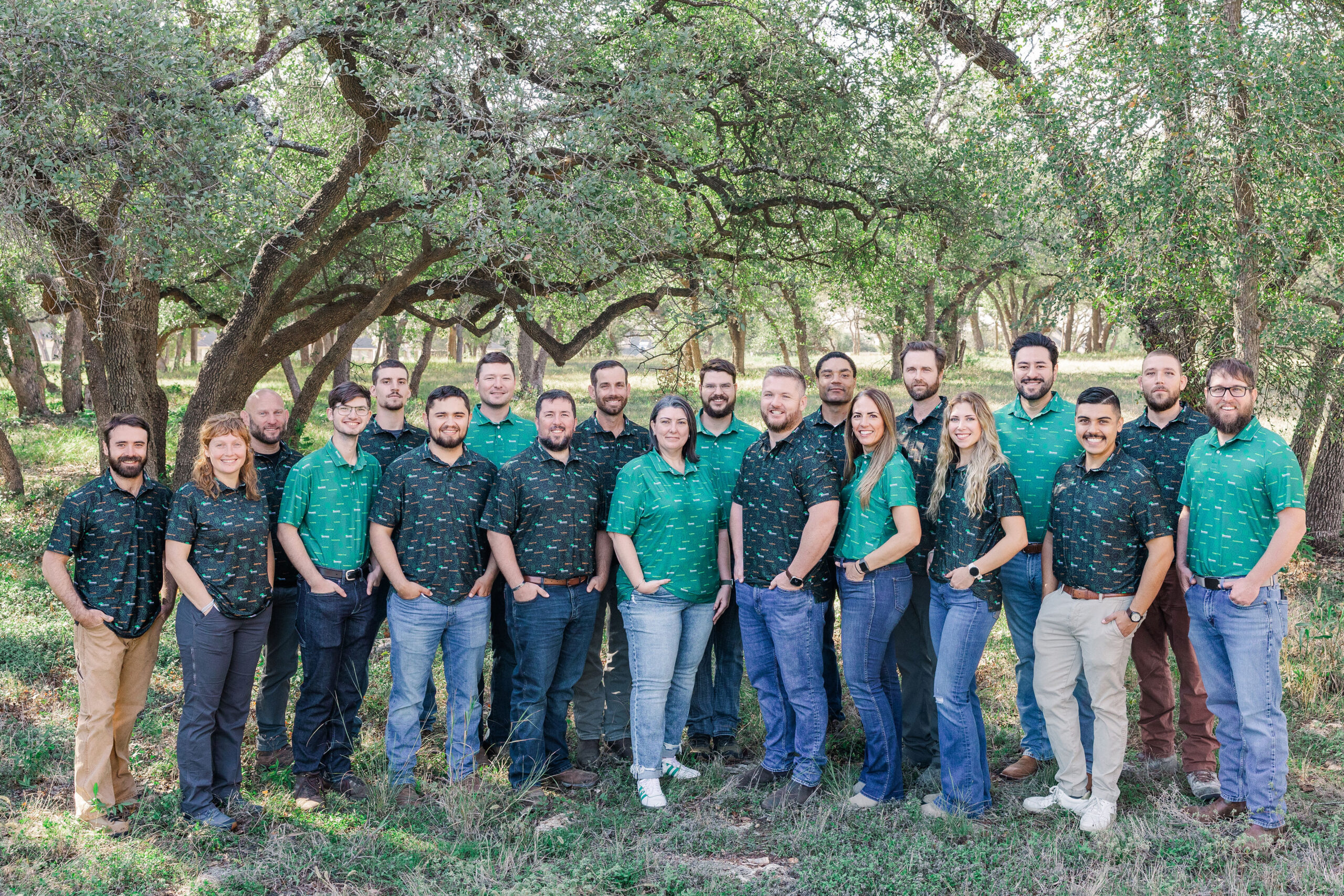 the Tree Mann Solutions team in coordinating green shirts smile at the camera in front of a canopy of trees