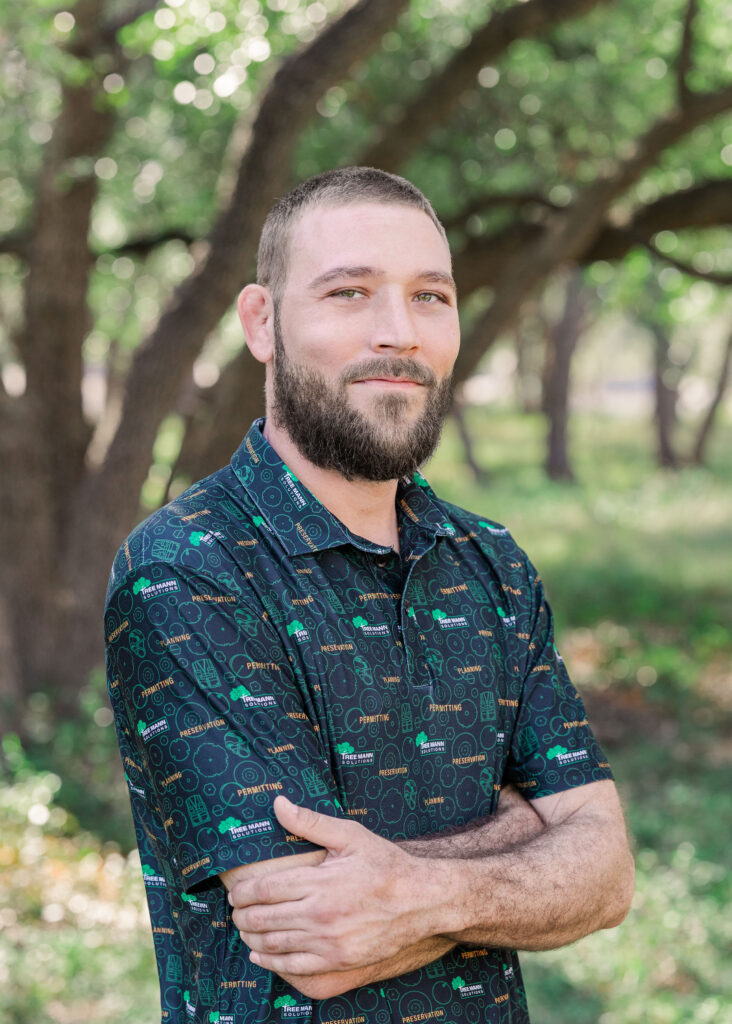 Cody Melton smiles at the camera with arms crossed wearing a dark green Tree Mann Solutions polo shirt with greenery in the background.