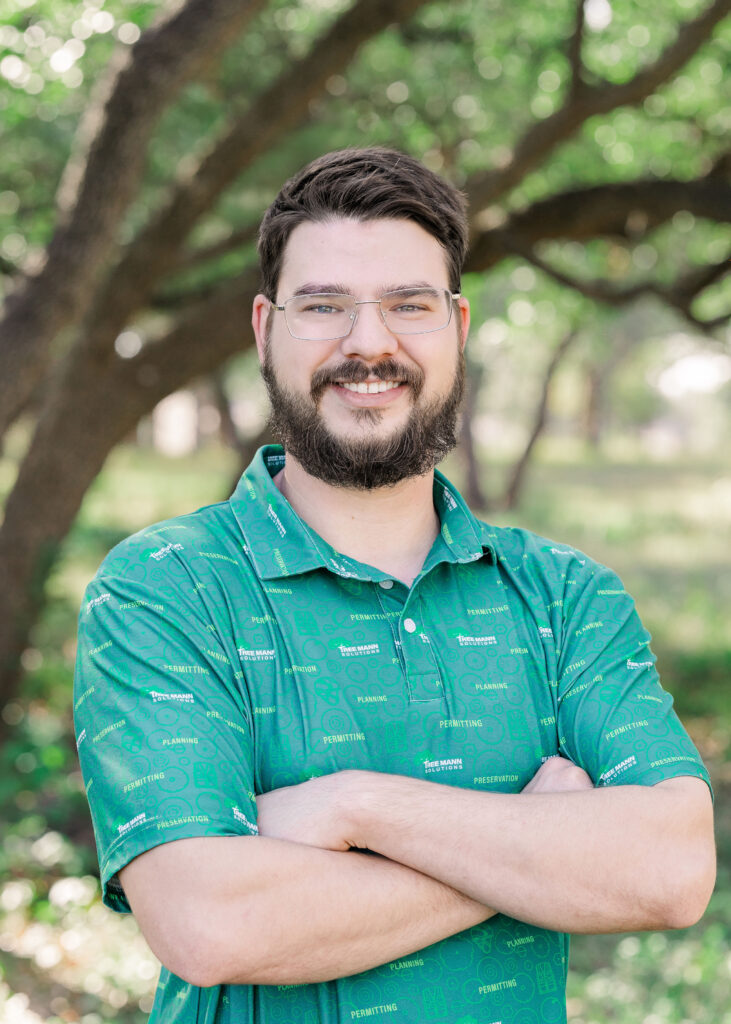 Micah Mabe smiles at the camera while crossing his arms and wearing a green shirt with trees in the background